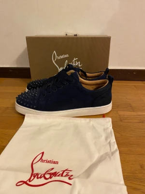 Mörkblå Christian Louboutin sneakers - Säljer ett par mörkblå sneakers från Christian Louboutin i mocka med ikoniska nitar på tån och vit sula. Skorna har klassisk röd undersida och svarta snören. Kommer med originalpåse och extra nitar. Riktigt snygga och unika sneakers för dig som vill sticka ut.