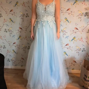 Ljusblå långklänning med spets och öppen rygg - Säljer en ljusblå långklänning med vacker spets upptill och glittriga detaljer. Klänningen har en öppen rygg med snörning och tunna axelband som korsas. Kjolen är i flera lager av tunn tyll för en drömmig look. Perfekt för bal!💕                          Aldrig använd!💕