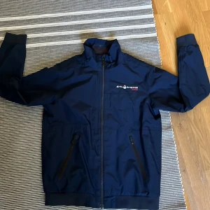 Marinblå Sail Racing Gore-Tex jacka - storlek 160 - Marinblå vindjacka från Sail Racing med Gore-Tex-material. Jackan har hög krage, dragkedja framtill och två fickor med dragkedja. Broderad logga på bröstet och ryggen. Perfekt för dig som gillar stilren och sportig look. Jackan är i strl 160. Skriv gärna om du har frågor!🤗.