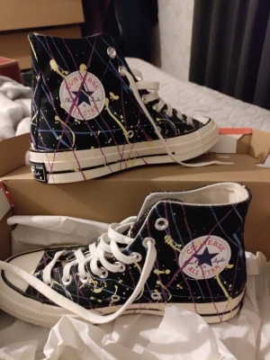 Converse All Star high top Chuck 70 HI 39,5 - Säljer ett par premium Converse All Star high top sneakers i svart med färgstänk och rutigt mönster i lila, rosa och gult. Klassisk rund tå, vita snören och sula. Ikonisk logga på sidan och canvasmaterial. Perfekta för dig som vill sticka ut med din stil. De är nästan nya inte mycket använda. Lådan är med.  Använda en eller två gånger bara