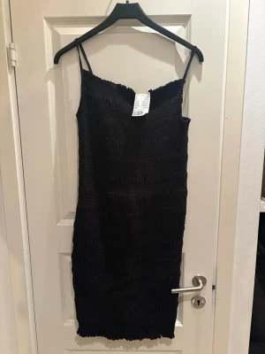 Svart smockad klänning H&M - Svart smal axelbandsklänning från H&M med smockad struktur. Klänningen är figurnära och har en enkel, stilren design med tunna axelband. Perfekt för dig som gillar minimalistisk stil och vill ha något lätt att matcha.