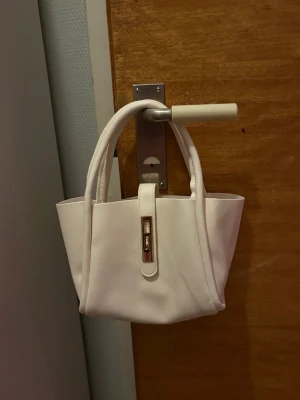 Vit handväska i skinn med gulddetalj - Snygg vit handväska i skinn med två handtag och en guldfärgad detalj framtill. Väskan har en stilren och enkel design som passar till många olika outfits. Perfekt storlek för det viktigaste du behöver ha med dig. DEFEKTER FINNS! SE SISTA BILDERNA! INGÅR ÄVEN EN LITEN NESSESÄR OCH EN RÄM TILL VÄSKAN OCH INGA DEFEKTER PÅ DOM!🥰❤️
