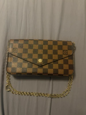 Rutig brun kuvertväska med kedja - Säljer en klassisk kuvertväska  i brunt och mörkbrunt rutigt mönster. Väskan har guldfärgad kedjerem och knappstängning framtill. Perfekt storlek för det viktigaste och ger en lyxig touch till din outfit.