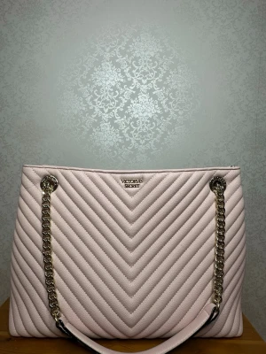 Ljusrosa quiltad axelväska Victoria's Secret - Snygg ljusrosa axelväska från Victoria's Secret med quiltad design i chevronmönster. Väskan har guldfärgade kedjeaxelband och en stilren metallogo framtill. Perfekt för dig som vill ha en trendig och rymlig väska med exklusiv känsla.