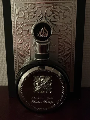 Lattafa Fakhar - Exklusiv parfym från Lattafa i en rund, svart flaska med eleganta silverdetaljer och orientaliskt mönster. Flaskan har en dekorativ silverkork och reliefmönster framtill. Perfekt för dig som gillar lyxig design och orientaliska dofter. Priset är ej hugget i sten!!!!