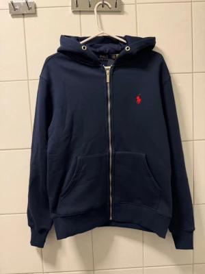 Marinblå hoodie från Polo Ralph Lauren - Snygg marinblå hoodie från Polo Ralph Lauren med dragkedja framtill och klassisk röd logga broderad på bröstet. Hoodien har huva med vita dragsnören och stora fickor framtill. Tillverkad i mjuk bomullsblandning för skön känsla.