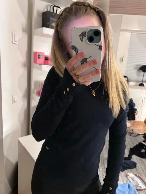 Svart långärmad tröja från holly&white - Snygg svart långärmad tröja från holly&white med rund halsringning och guldknappar vid ärmsluten. Tröjan har en figurnära passform och diskret logga vid höften. Perfekt för en stilren look.