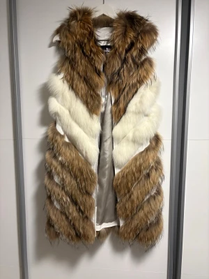 Pälsväst från Gold Frost i brunt och vitt - Snygg pälsväst från Gold Frost i storlek M. Västen har ett mönster i diagonala ränder med bruna och vita partier, och är tillverkad i fuskpäls med silkeslen insida. Perfekt för att lyfta din outfit med en lyxig touch.