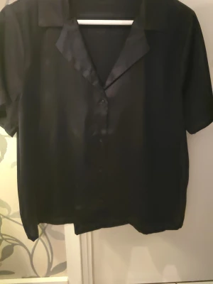 Svart kortärmad skjorta i satin - Stilren svart skjorta med korta ärmar och klassisk krage. Skjortan är tillverkad i ett glansigt satinliknande material och har knäppning framtill. Perfekt för dig som gillar en enkel men trendig look.