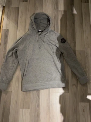 Grå hoodie med patch på ärmen - Snygg grå hoodie med huva och dragsnören. Klassisk känguruficka framtill och en rund svart patch på vänster ärm som detalj. Tillverkad i mjukt sweatshirtmaterial, perfekt för chill dagar. Enkel och stilren design som funkar till det mesta pris kan diskuteras .