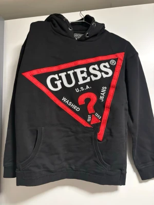 Svart hoodie från Guess med stor logga - Svart hoodie från Guess med stor, ikonisk röd och vit trekantig logga på bröstet. Klassisk huva med dragsko och magficka framtill. Tillverkad i mjukt material som passar perfekt för en avslappnad och trendig stil.