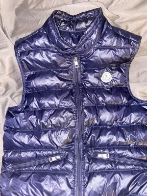 Mörkblå dunväst från Moncler - Snygg mörkblå dunväst från Moncler med glansig finish och ståkrage. Västen har två dragkedjeförsedda fickor framtill och klassisk Moncler-logga på bröstet. Perfekt för lager-på-lager och håller dig varm med stil. Använd två gånger och säljer för den är för liten. Det är barn storlek o passar mellan 158-164. Kan gå ner i pris till 1800.ÄKTA