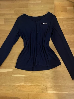 Mörkblå ribbad kofta från Levi's - Mörkblå ribbad kofta från Levi's med lång ärm och knäppning framtill. Tröjan har en figurnära passform och diskret logga på bröstet. Perfekt för lager-på-lager och enkel att matcha med jeans eller kjol.