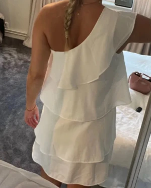 Vit one shoulder volangklänning - Säljer en superfin vit klänning med one shoulder och flera lager volanger. Klänningen är kort och har en luftig passform som känns perfekt för sommardagar. Materialet är lätt och känns som polyester eller liknande. Riktigt snygg och trendig modell!