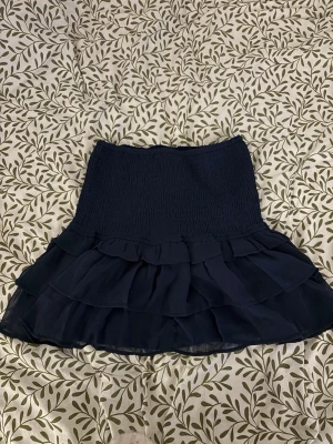 Volangkjol från Gina Tricot, XS - Mörkblå kortkjol med smockad resårmidja med volanger från Gina Tricot. Kjolen har lager med volanger med stretchigt midja . Materialet känns lätt och luftigt. Är använd ett få tal gånger, men inga tecken på användning. 