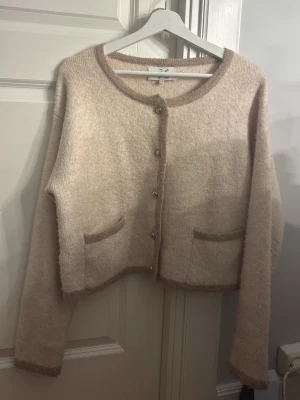 Beige kofta med knappar och fickor - Mysig beige kofta med rund halsringning, dekorativa guldknappar och två framfickor. Koftan har en lös passform och är gjord i ett mjukt, stickat material med kontrasterande kanter.