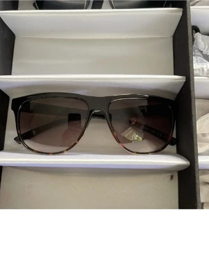 Svarta VonZipper solglasögon - Snygga solglasögon från VonZipper med svart båge och gulddetaljer på sidorna. Glasen är tonade i brunt och har en rektangulär form som ger en cool och modern look. Perfekta för dig som vill sticka ut med stilrena accessoarer.