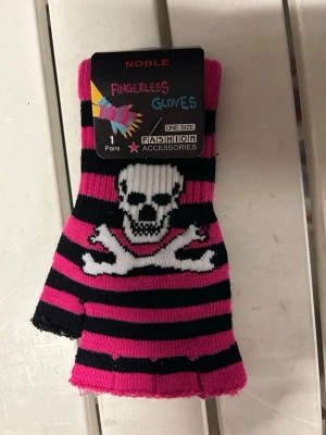 Vantar i onesize. Ceriserosa/svart randiga. - Fingerlös handske i svart och ceriserosa ränder med en vit dödskalle och korslagda ben framtill. Stickad i mjukt material och passar perfekt för en edgy stil. One size.