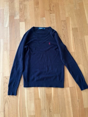 Polo Ralph Lauren - Svart stickad tröja från Polo Ralph Lauren med rund halsringning och lång ärm. Tröjan har en liten röd broderad logotyp på bröstet och ribbstickade muddar vid ärmslut och nederkant.  Tröjan har lite knottrer, går ju att få väck med sån maskin vissa har. Men tröjan är i fint skick för att vara en stickad tröja:)
