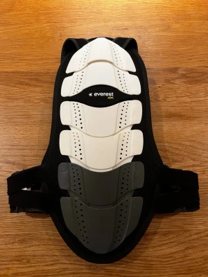 Ryggskydd Everest ADV svart/vit - Säljer ett ryggskydd från Everest ADV med hårda vita och grå skyddsplattor och svart meshmaterial. Baksidan har justerbara axelband, midjebälte med kardborre och gula detaljer. Perfekt för skidåkning, snowboard eller cykling.