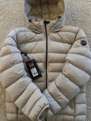 Colmar jacka - Colmar Expert Jacket | Snygg sandfärgad/vit dunjacka från Colmar med huva som är perfekt för kallare väder. Storleken är 48 vilket morsvarar S i vanliga plagg, (jag är 175 på bilden). Skick 8/10. Nyligen tvättad hos tvätteri. Ett minimalt hål på höger underarmen som inte läggs märke till (som du märker på övriga bilderna), men är täckt med en liten reflex för att den inte ska tappa dun. Digitalt kvitto finns! Samt QR kod och lappar för jackan. 