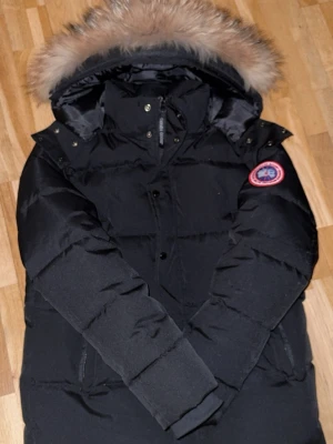 Svart dunjacka från Canada Goose - Snygg svart dunjacka från Canada Goose med klassisk patch på ärmen och stor huva med fluffig päls. Jackan har långa ärmar, dragkedja och knappar framtill samt två fickor. Perfekt för kalla vinterdagar och riktigt varm.