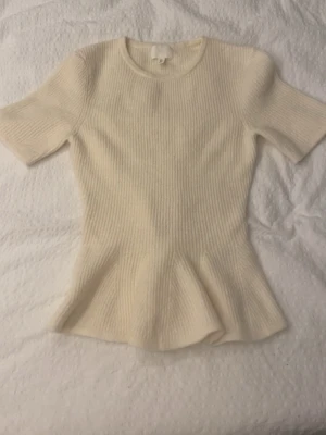 Ribbad cremefärgad topp från H&M - Säljer en ribbad cremefärgad topp från H&M med korta ärmar och lätt utsvängd nederkant. Toppen har rund halsringning och är gjord i ett mjukt stickat material som ger en bekväm och snygg passform.