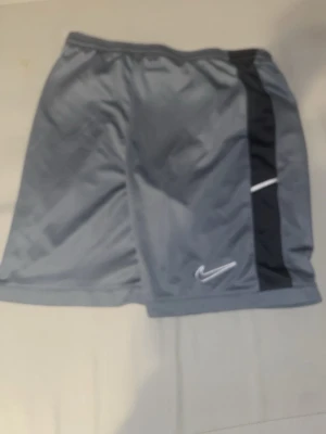 Grå sportshorts från Nike - Snygga grå sportshorts från Nike med svart sidopanel och vit logga. Perfekta för träning eller chill, med bekväm passform och elastisk midja. Lätta och luftiga i syntetmaterial som andas.