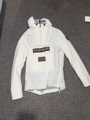 Vit vindjacka från Napapijri - Snygg vit vindjacka från Napapijri med huva, stor magficka med logga och norsk flagga på bröstet. Jackan har dragkedja på sidan och upptill, samt justerbar huva. Perfekt för blåsiga dagar och streetwear-stil.