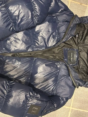 Mörkblå dunjacka från EX - Snygg mörkblå dunjacka från EX med quiltad design och dragkedja framtill. Jackan har en hög krage och en patch med EX-logga på ärmen. Perfekt för kalla dagar och har en modern, puffig look.