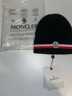 Svart Moncler mössa med logga - Snygg svart ribbstickad mössa från Moncler med klassisk logotyp framtill. Mössan har en rand i rött och vitt runt kanten som ger en sportig vibe. Perfekt accessoar för att lyfta din outfit under kyliga dagar.