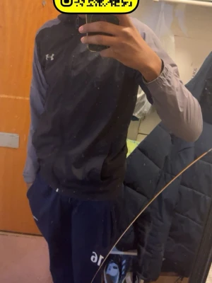 Svart och grå vindjacka Under Armour - Snygg svart och grå vindjacka från Under Armour med dragkedja framtill och långärmad design. Jackan har en sportig look och är tillverkad i lätt polyester, perfekt för aktiva dagar. Under Armour-logga på bröstet ger en clean och modern känsla.