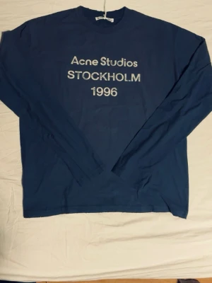 Blå långärmad tröja Acne Studios - Blå långärmad tröja från Acne Studios med vit text framtill: 'Acne Studios STOCKHOLM 1996'. Tröjan har rund halsringning och är tillverkad i mjuk bomull. Perfekt för en avslappnad och trendig look.