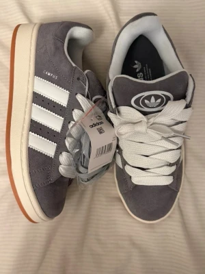 Grå adidas Campus sneakers mocka - Snygga grå adidas Campus sneakers i mocka med klassiska vita ränder på sidorna och vit sula med brun yttersula. Skorna har rund tå, vita skosnören och adidas-logga på plösen. OBS tyvärr finns ingen skor box med