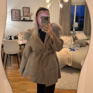 Beige kappa med päls  - Snygg beige kappa med stor huva kantad av päls. Kappan har knytskärp i midjan och vida ärmar för en trendig look. Perfekt för kyliga dagar och ger en lyxig känsla till outfiten. Furstockholm