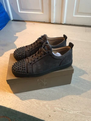 Grå mockasneakers med nitar från Louboutin - Säljer ett par grå sneakers från Christian Louboutin i mocka med svarta nitar på tån och klassisk röd sula. Skorna har snörning och en rund tå, vilket ger en edgy och lyxig vibe. Perfekta för dig som vill sticka ut med exklusiva detaljer.