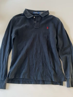 Svart långärmad polotopp Ralph Lauren - Klassisk svart långärmad polotopp från Ralph Lauren i slim fit-modell. Toppen har krage, knappar framtill och den ikoniska röda logotypen broderad på bröstet. Tillverkad i mjuk bomull som känns skön mot huden.
