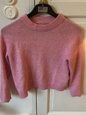 Rosa stickad tröja med bred krage - Mysig rosa stickad tröja med bred ribbad krage och långa ärmar. Tröjan har en rak, något kortare passform och ribbade muddar vid ärmslut och nederkant. Perfekt för lager på lager och enkel att matcha till jeans eller kjol.