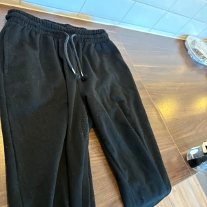 Svarta mjukisbyxor med snörning - Svarta mjukisbyxor med elastisk midja och snörning. Klassisk passform med muddar vid bensluten. Perfekta för chill eller träning. Tillverkade i mjukt material som känns skönt mot huden.