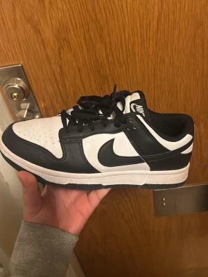Nike Dunk Low svart/vit sneaker - Klassiska Nike Dunk Low sneakers i svart och vitt med rund tå och snörning. Skon har en låg siluett, vit ovandel i läder med svarta overlays och svart Swoosh. Perfekt för dig som gillar stilrena och tidlösa sneakers.