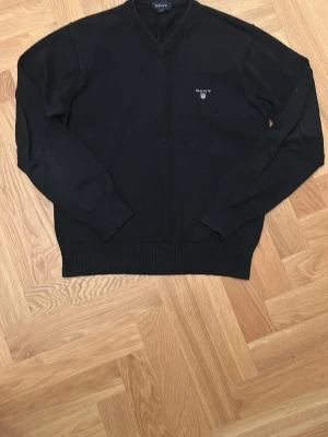 Svart stickad tröja från Gant - Klassisk svart stickad tröja från Gant med V-ringning och diskret logga broderad på bröstet. Tröjan har långa ärmar, ribbade muddar och nederkant. Perfekt till lager-på-lager och enkel att matcha med allt.