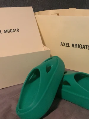 Gröna Axel Arigato tofflor - Säljer ett par gröna tofflor från Axel Arigato. De har en chunky, modern design med rund tå perfekta för dig som gillar stilrena och bekväma skor. Box och påse från Axel Arigato medföljer. 