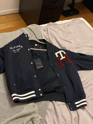 Mörkblå bomberjacka Tommy Hilfiger - Snygg mörkblå bomberjacka från Tommy Hilfiger med vita och vinröda detaljer. Jackan har broderad logga på bröstet, stora bokstäver 'TH' och klassiska ränder på muddar och krage. Perfekt för dig som vill ha en sportig och trendig look.