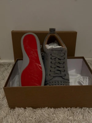 Gråa Louboutin sneakers med nitar - Snygga grå sneakers från Christian Louboutin med ikonisk röd sula och coola nitar framtill. Skorna har snörning och är gjorda i mocka med detaljer i skinn. Perfekta för dig som vill sticka ut med en lyxig och edgy stil.