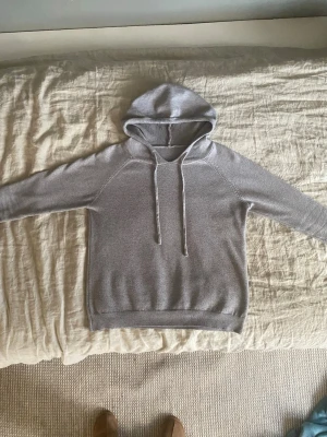 Ljusgrå hoodie med dragsko - En stilren ljusgrå hoodie med huva och långa ärmar. Tröjan har dragsko i huvan och ribbade muddar vid ärmslut och nederkant. Tillverkad i mjukt stickat material som känns skönt mot huden. Perfekt för en avslappnad look.