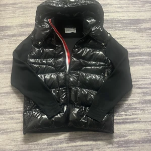 Svart dunväst kardigan från Moncler XS/S/M/ - Säljer en svart och mörk blå dunväst kardigan från Moncler med glansig yta och stickade svarta ärmar. Västen har dragkedja med röd och vit detalj längs öppningen samt två fickor med dragkedja. Klassisk Moncler-logga på ärmen och avtagbar huva på vissa bilder.