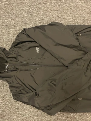 Svart vindjacka från New Balance M - Svart vindjacka från New Balance med huva och hel dragkedja framtill. Jackan har två sidofickor med dragkedja och justerbar dragsko vid huvan. Perfekt för blåsiga dagar och sportiga outfits.