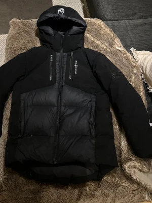 Svart dunjacka från Sail Racing - Storlek 150 den motsvarar (XS) Säljer en svart dunjacka från Sail Racing med huva och två bröstfickor med dragkedja. Jackan har Sail Racing-logga på bröstet och är fylld med Allied Feather + Down (500 FP). Perfekt för kalla dagar och riktigt snygg streetstil. Köpt för 3200kr!,, det är barnstorlek