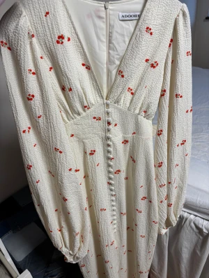 Vit blommig långklänning från Adoore - Superfin långklänning från Adoore i vit crepad bomull med små röda blommor. Klänningen har v-ringning, långa puffärmar och knappar framtill. Perfekt för sommarens alla tillfällen och ger en romantisk vibe. Dåligt ljus på bilden tyvärr men jättefint skick! Använd en gång! 1300kr