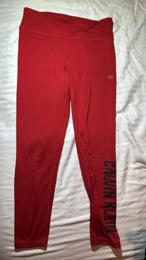 Röda leggings från Calvin Klein - Snygga röda leggings från Calvin Klein med hög midja och svart logotyp längs benet. Perfekta för en sportig look eller chill outfit. Mjukt och stretchigt material som sitter tight och bekvämt.